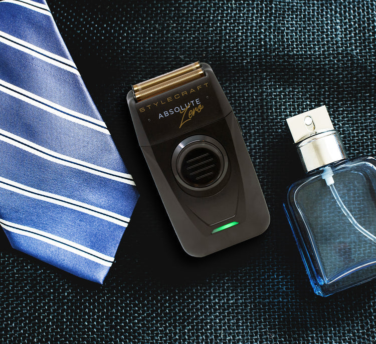 A black and gold Stylecraft Absolute Leo electric beard trimmer lies on a dark textured surface, styled with a blue and white striped necktie on the left and a clear perfume bottle on the right.