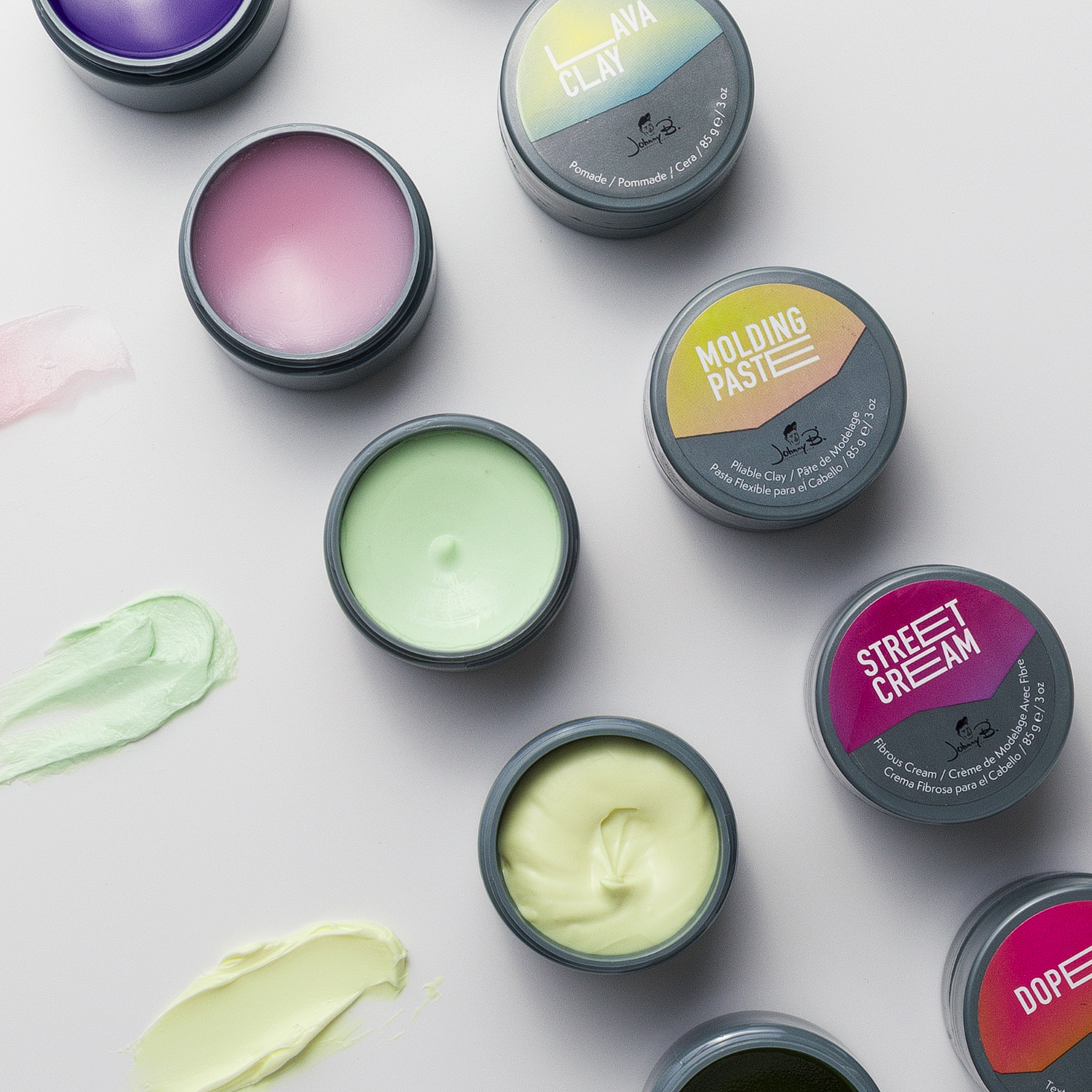 An overhead flat-lay of several open jars of clay makeup and colored cream pastes on a light gray background, with matching cream swatches smeared beside them. Visible product names include Clay Clay, molding paste, Street Cream, and Dope.