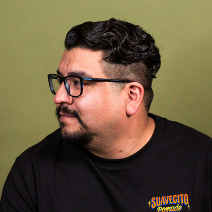 A man with dark hair, a beard, and glasses wearing a black t-shirt with a colorful graphic on the left chest that reads 'SUAVECITO Pomade'. He is photographed in profile against a solid olive green background.