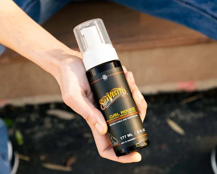 Hand holding a Suavecito Curl Mousse bottle with a blurred outdoor background